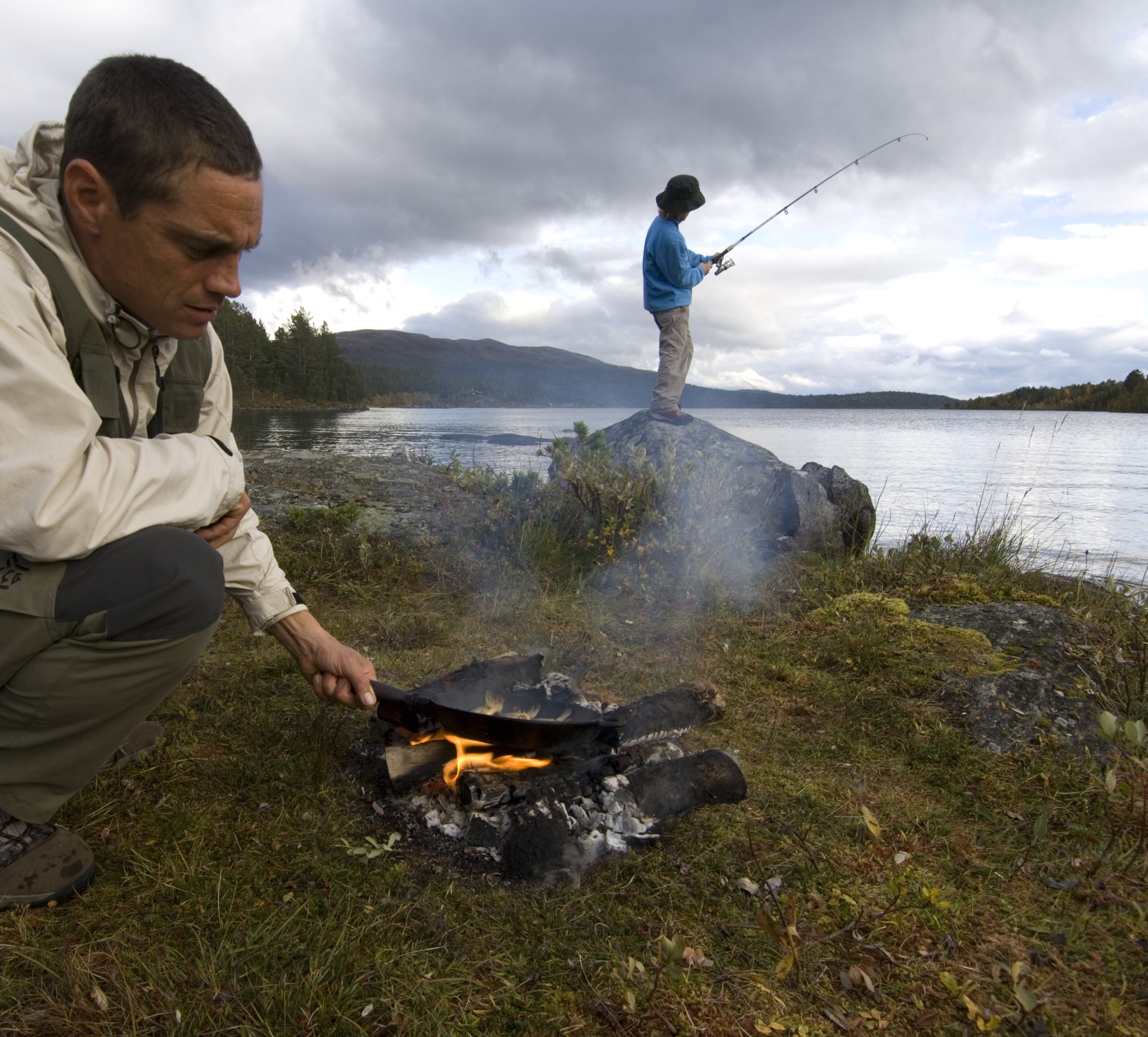 Two people by a mountain lake with fishing rods and a campfire