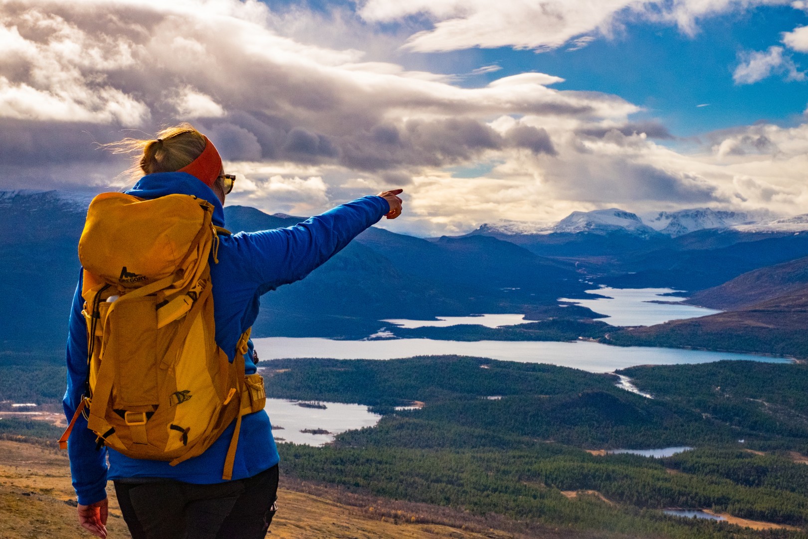 Person standing on a hill pointing over a landscape with mountain lakes and mountains